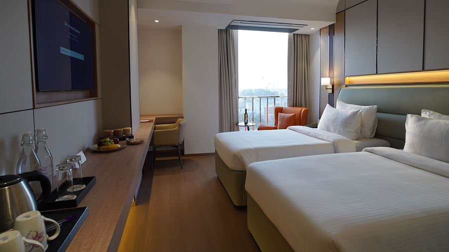 A modern hotel room includes two single beds with white linens, a seating area with a dark sofa, and a view towards a window with city glimpses - The Orchid Hotel, Chandigarh