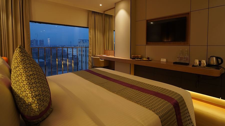 A stylish hotel room showcases a bed with striped bedding facing a large window with city views at dusk, a TV, and a desk - The Orchid Hotel, Chandigarh