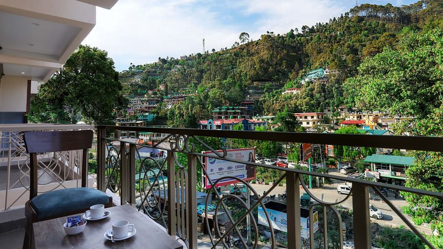 A building's balcony adorned with a chair, a coffee table bearing two tea cups and a bowl, all with a view overlooking a picturesque mountain town - Vinsober Monal, Nainital