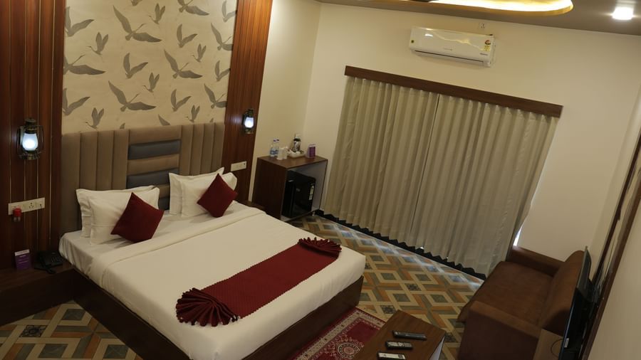 An overview of an Executive Room at VITS Select Diamond Resort, Sasan Gir. The image shows a bed, a small sofa, a ceiling fan, and an air conditioner with a view of curtains and a window.