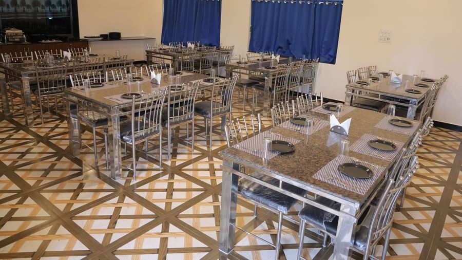 A dining area with multiple tables and chairs, each set with plates, cutlery, and glasses. The tables are granite with steel frames, and there is a buffet counter in the background.