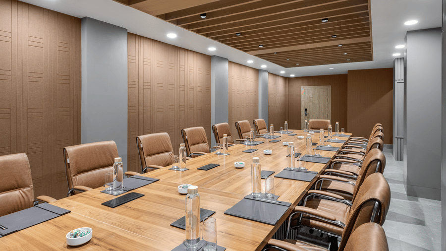A conference room featuring a long, light wood meeting table surrounded by brown upholstered chairs, with wood-panelled walls and a matching suspended ceiling section | Voyage Eastin Suites