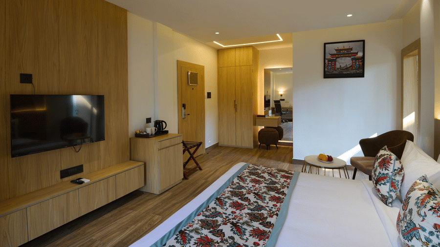 A wide shot of a hotel suite or large room with a double bed, a long patterned floor runner, light wood panelling, and a seating area | Voyage Eastin Suites