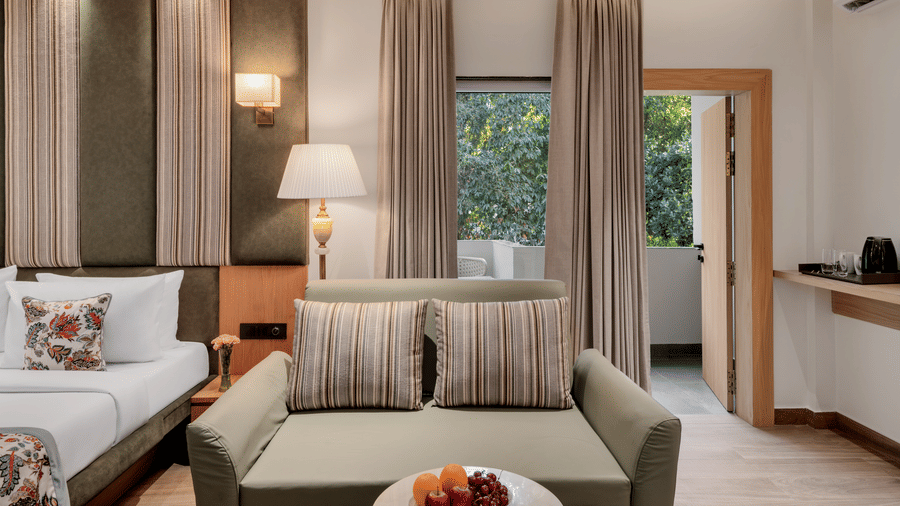 A hotel suite with a sofa seating area near the glass balcony doors, a large bed with floral bedding, and dark wood panelling and curtains | Voyage Eastin Suites