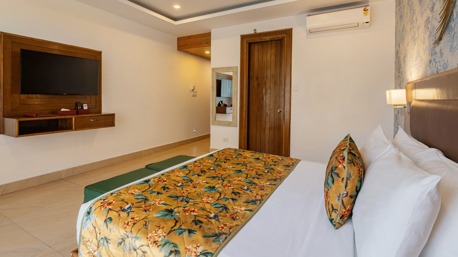 A room with an attached bathroom, featuring a bed, a television unit, a small wall mirror, a decorative ceiling and tiled floor | Summit by the Ganges Beach Resort & Spa, Rishikesh