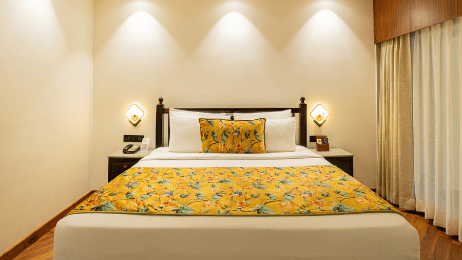 A bedroom with a wooden bed,  2 small cabinets, 2 bedside lamps, and opaque and sheer curtains | Summit by the Ganges Beach Resort & Spa, Rishikesh