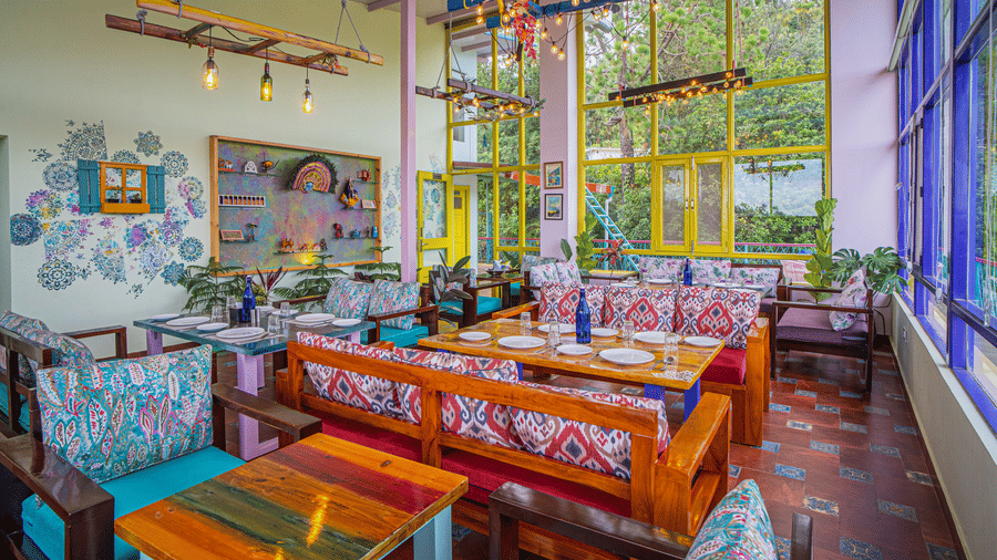 A dining area with tables, chairs, large windows, and decor items arranged indoors at Adrushya Estates, Mukteshwar.