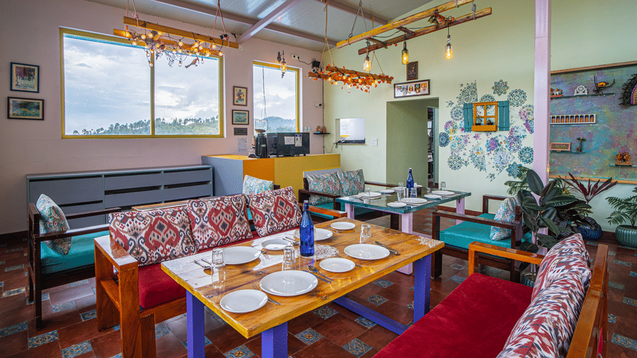 An indoor dining area with multiple tables set with plates, sofas, wall decor, and large windows at Adrushya Estates, Mukteshwar.