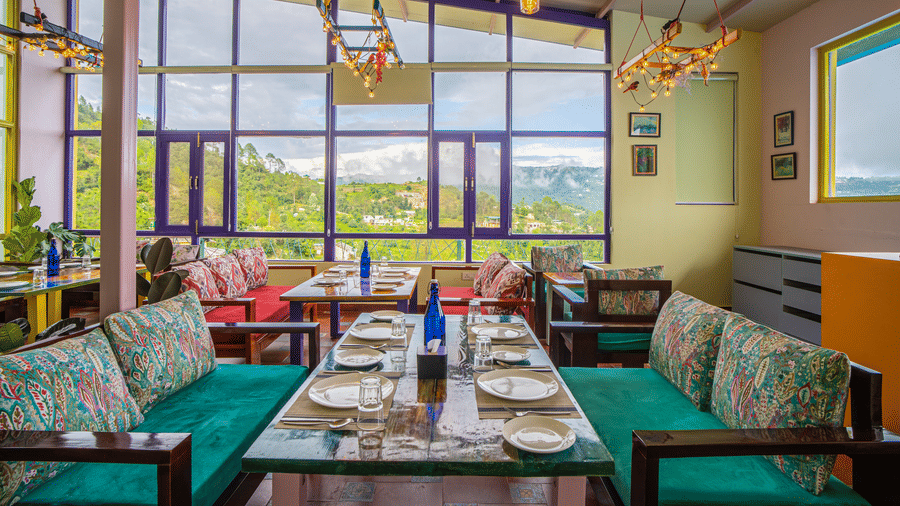A dining area with tables, cushioned benches, hanging lights, and large glass windows at Adrushya Estates, Mukteshwar.