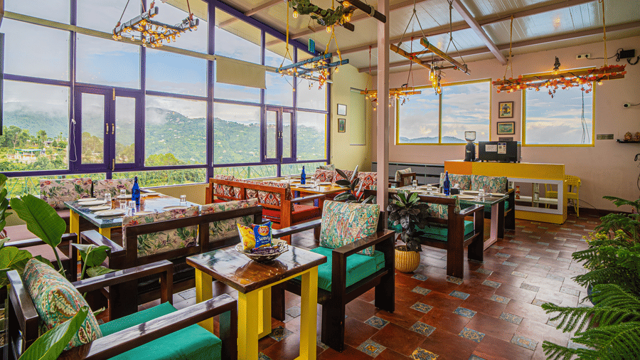 An indoor dining space with tables, chairs, plants, and large windows overlooking hills at Buransh by Adrushya, the best restaurant in Mukteshwar