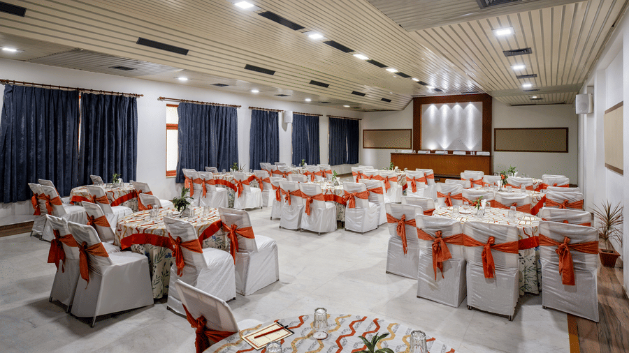 A large banquet hall featuring multiple round tables covered in white tablecloths with red ribbons on chairs at Amantra Shilpi Resort, Udaipur.