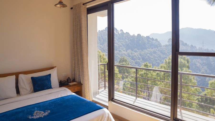 A view from inside the Sun Terrace Room at BluSalzz Escapade Resort, Lansdowne, shows a bed on the left and a large glass window structure on the right. The windows look out onto a hillside covered in trees.