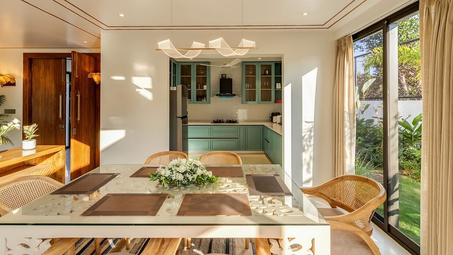 A bright dining area with a large wooden table and chairs, overlooking an open kitchen and outdoor greenery - BluSalzz Villas - Alocassia, Anjuna