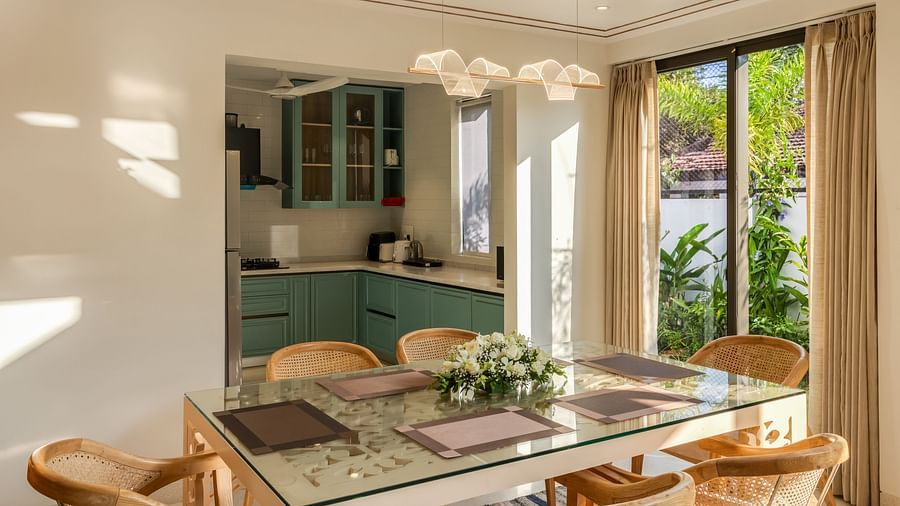 A dining area with a modern wooden table and chairs, adjacent to a bright kitchen with a large window offering a lush outdoor view.
