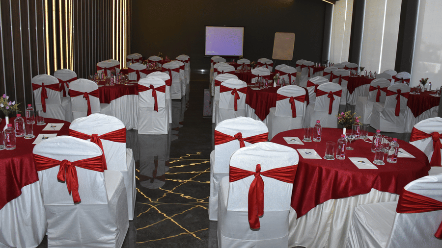 A banquet hall at Comfort Inn Sapphire by Eastlynn Collections, Jaipur with round tables, chairs tied with red sashes, and stage setup.