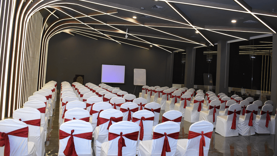 A banquet hall at Comfort Inn Sapphire by Eastlynn Collections, Jaipur with arranged seating, stage screen, and patterned ceiling lights.
