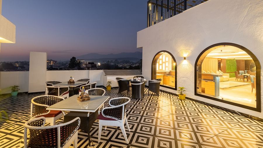 Image of the rooftop restaurant at DLS Grand Luxotica, Dehradun during evening featuring chairs and tables arranged on a well-furnished floor