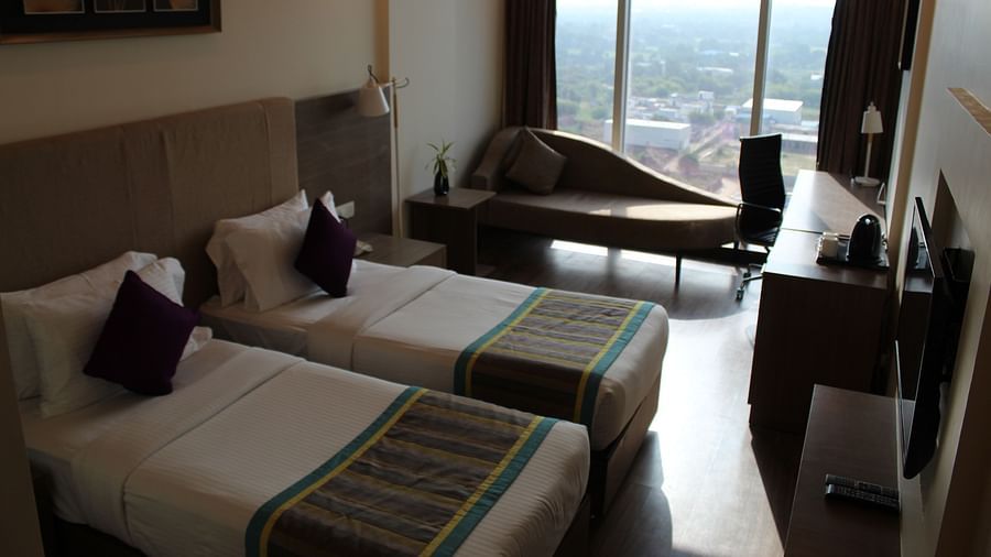 An dim-lit room showcasing a pair of twin beds with lush decors, a long sofa with cushion, and a flat screen TV, inside a room with open curtains.