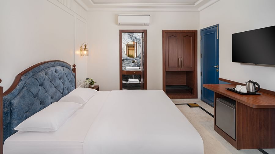 A side view of the room features a spacious bed, a wall-mounted television directly opposite the bed, and wooden furniture | Hotel Ganga Maiya