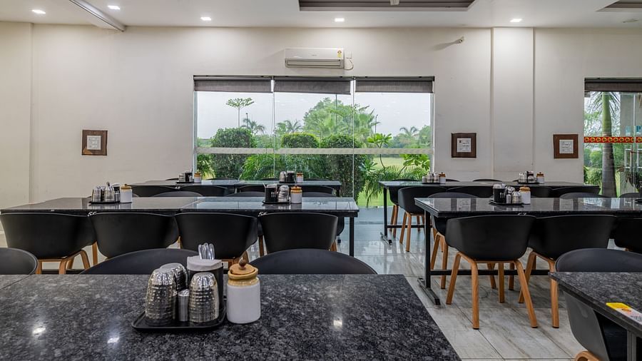Front view of the dining tables reflecting the overhead lights, and glass windows offering view of the trees outdoor at Hotel Highway King, Behror.