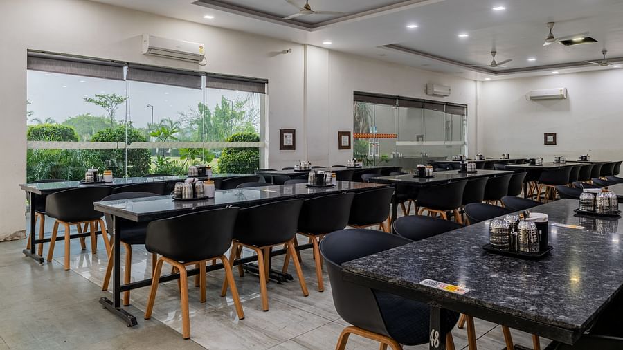 Air-conditioned restaurant of Hotel Highway King, Behror, is furnished with parallel aligned dining tables, water cups and glass windows that offer views of the trees outside at Hotel Highway King, Behror.