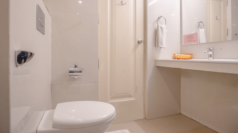 A simple, white-themed restroom with a wall-mounted toilet, a sink, and a plain door at Hotel Horizon, Trivandrum.