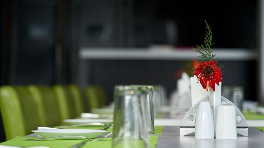 A close-up of a table setting in the restaurant at Hotel Mamallaa Heritage, showing a white plate, glassware, and a small red floral arrangement on a green placemat.