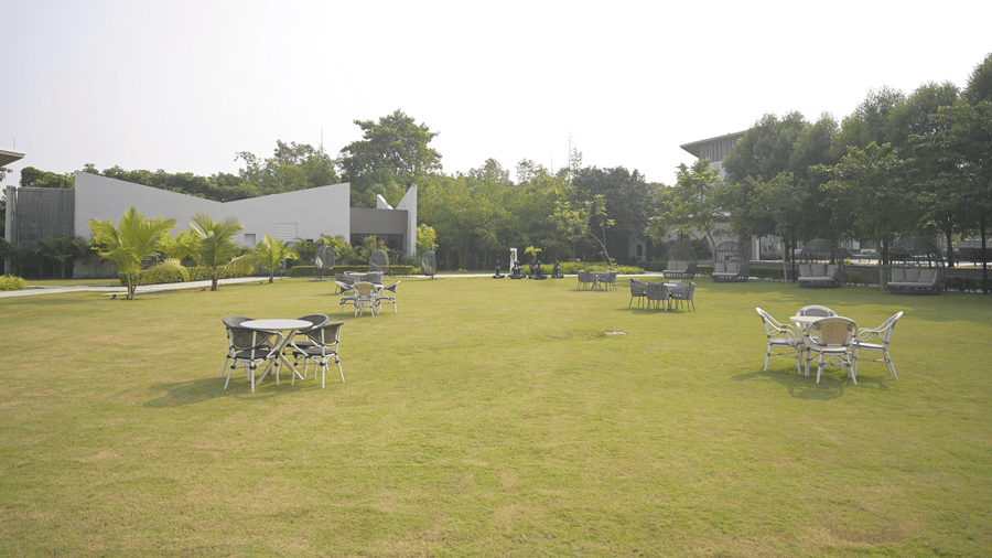 A wide green garden with scattered outdoor tables and chairs surrounded by lush plants at Hotel Sonar Bangla, Mayapur.