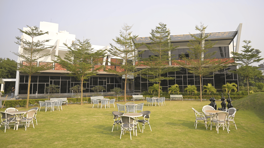 A green lawn with several sets of outdoor tables and chairs in front of a glass building at Hotel Sonar Bangla, Mayapur.
