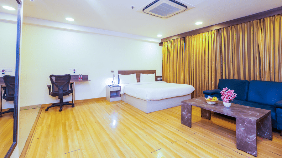 A bright hotel room with a large bed, wooden floors, a blue sofa, and a desk with a chair - Hyphen Business Hotel, Noida