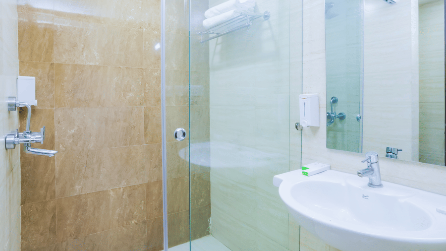 Well-lit bathroom featuring a glass-enclosed shower area, sink, and warm beige tiling at Hyphen Premier Hotel, Meerut
