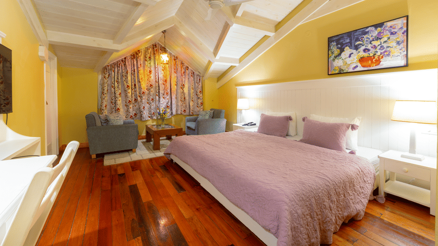 A bright attic-style bedroom featuring a bed with purple linens, vaulted white ceilings, and a small sitting area - Johnson Lodge & Spa, Manali