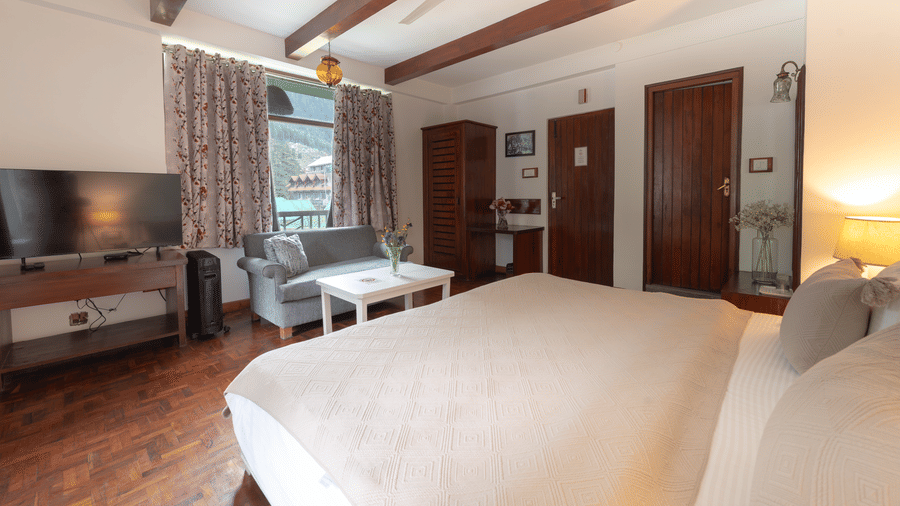 Perspective of a guest suite highlighting the dark wood ceiling beams, wall-mounted TV, and bright interiors - Johnson Lodge & Spa, Manali