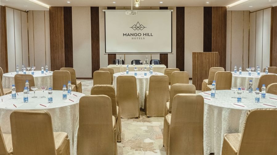 Banquet hall at Mango Hill Central, Chennai, with several round tables and chairs. A projector screen with the hotel logo is at the front of the room.