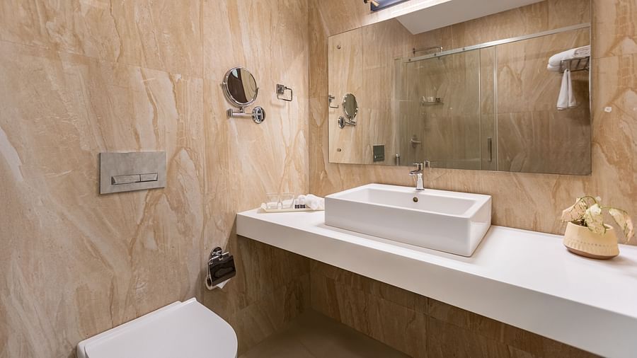 A bathroom with beige and brown textured walls, a white rectangular sink, a large mirror, and a white toilet - Mango Hill Resort, Coimbatore