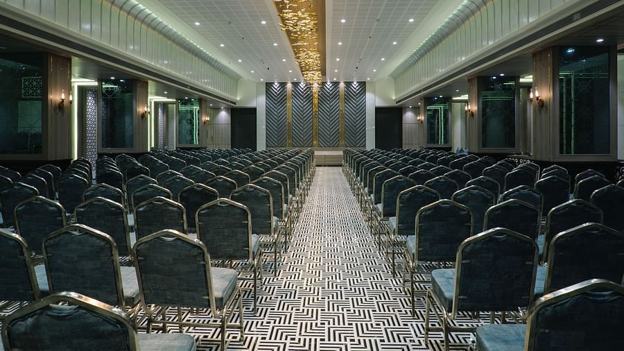 A large room is set up with rows of dark chairs facing a stage or presentation area - Melior Inn