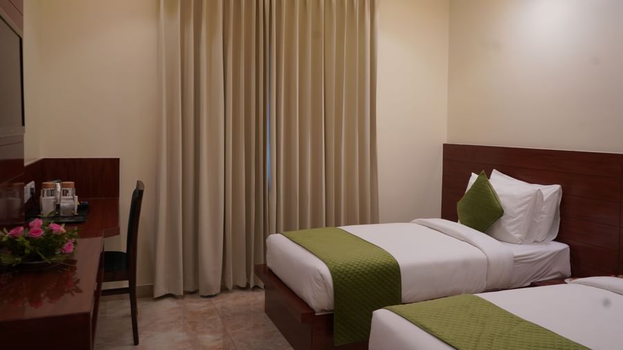 A wide shot of a hotel room with two single beds. The beds have white comforters with green accents and are separated by a small table.