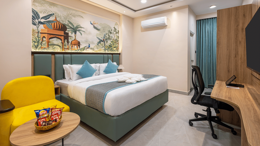 A bedroom with a king size bed with white linens, a yellow armchair, small table with snacks, a wall-mounted television, and a work desk  with a chair, at Monday Hotels & Suites Madhapur Hyderabad.