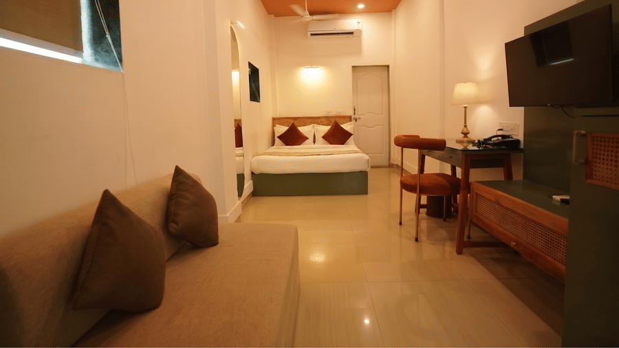 A bedroom at Oceanature Resort, Alibaug with a sofa, awall-mounted television, a wooden desk with chairs, and a queen bed in the background.