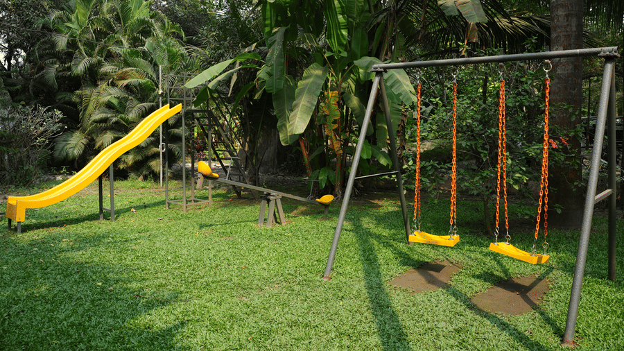 A children's playground area at Oceanature Resort, Alibaug features a yellow slide, a see-saw, and swings on a green lawn.