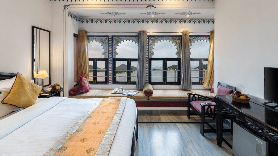 A side view of the bedroom featuring a spacious bed, a wooden table under the wall-mounted television, and bay window seating with curtains pulled to the side at Panna Vilas Palace