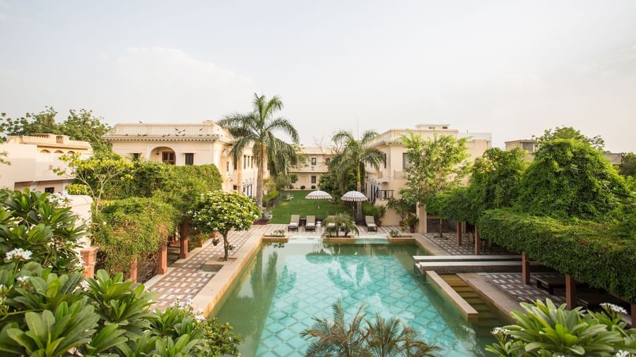 A serene view of an outdoor swimming pool at Royal Heritage Haveli surrounded by green trees and buildings under a clear sky.