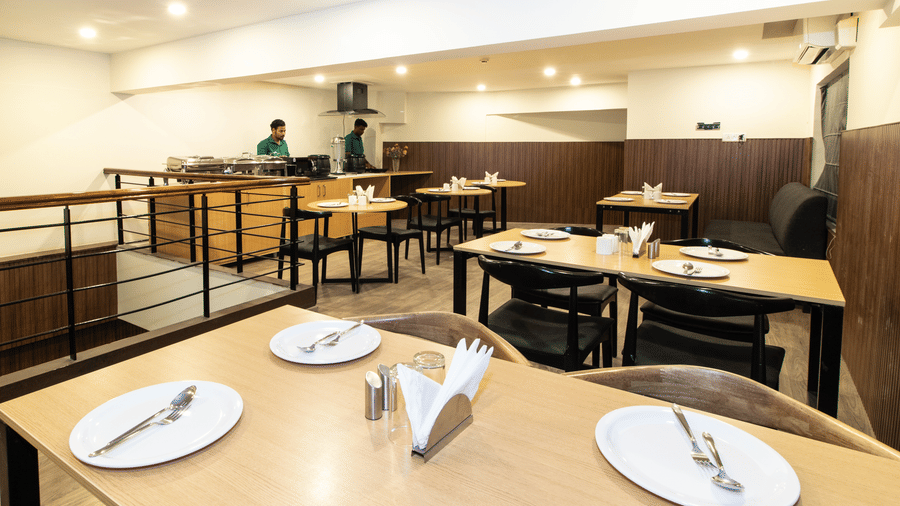 The restaurant at Sanctum Suites, Richmond Road, Bangalore shows wooden tables set with white plates, cutlery, black chairs, a railing, and a buffet counter.