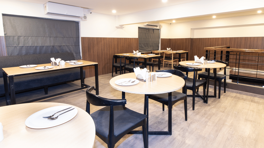 Sanctum Suites, Richmond Road, Bangalore features a dining room with circular wooden tables, black chairs, a window with a dark blind, and ceiling lights.
