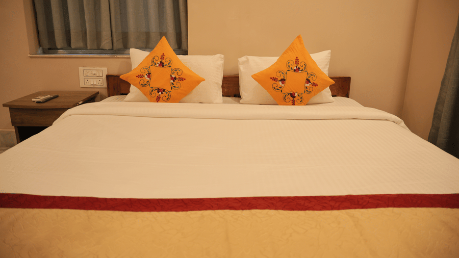 A close-up view of a king size bed featuring bed sheets, a runner, and two bright yellow accent pillows, with a side table, at Shophori Wellness Retreat, Bolpur.