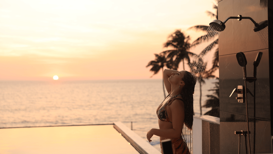 A person standing by an outdoor pool and shower fixture at sunset at SUKAVI, facing a vast ocean with a few palm trees visible in the background.
