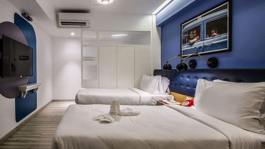 A spacious and well-lit hotel room with twin beds, a TV mounted on the wall, and a large mirror  | T24 Retro