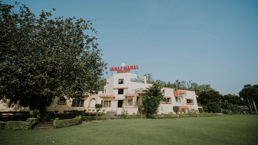 A facade view across a large green lawn looking towards the building exterior of TGI Jahaj Mahal, Mandu, partially framed by trees.