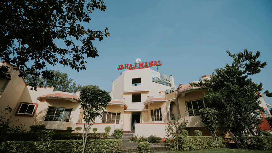 An exterior facade of TGI Jahaj Mahal, Mandu, surrounded by green trees and bushes under a clear sky.