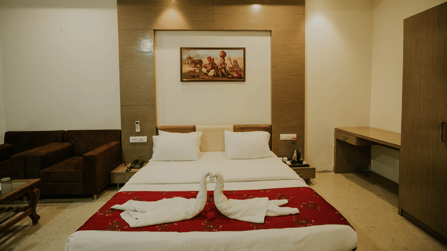 A bedroom at TGI Jahaj Mahal, Mandu, features a king size bed with white linens and towel art, a sofa, and a framed picture on the wood-panelled wall.